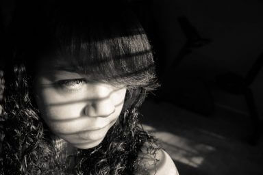 A somber looking girl with bangs stares out the window as a representation of the signs of depression