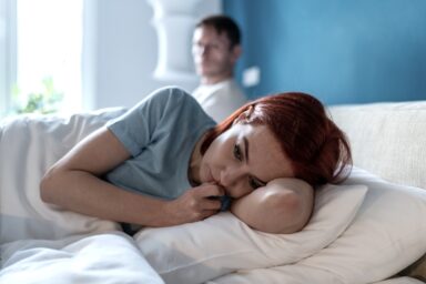 Sad woman lies awake in bed while her partner looks on in the background